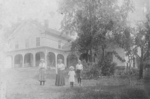 Historic, black and white photo of two women and two children standing in front of a white farmhouse in Chatham, New York.